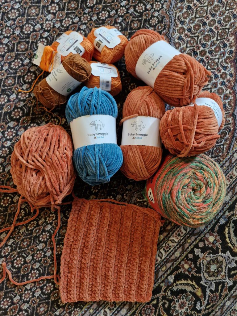 Skeins of Hobbii Baby Snuggle velvet yarn in orange and wintergreem with a ribbed crochet swatch on a patterned rug