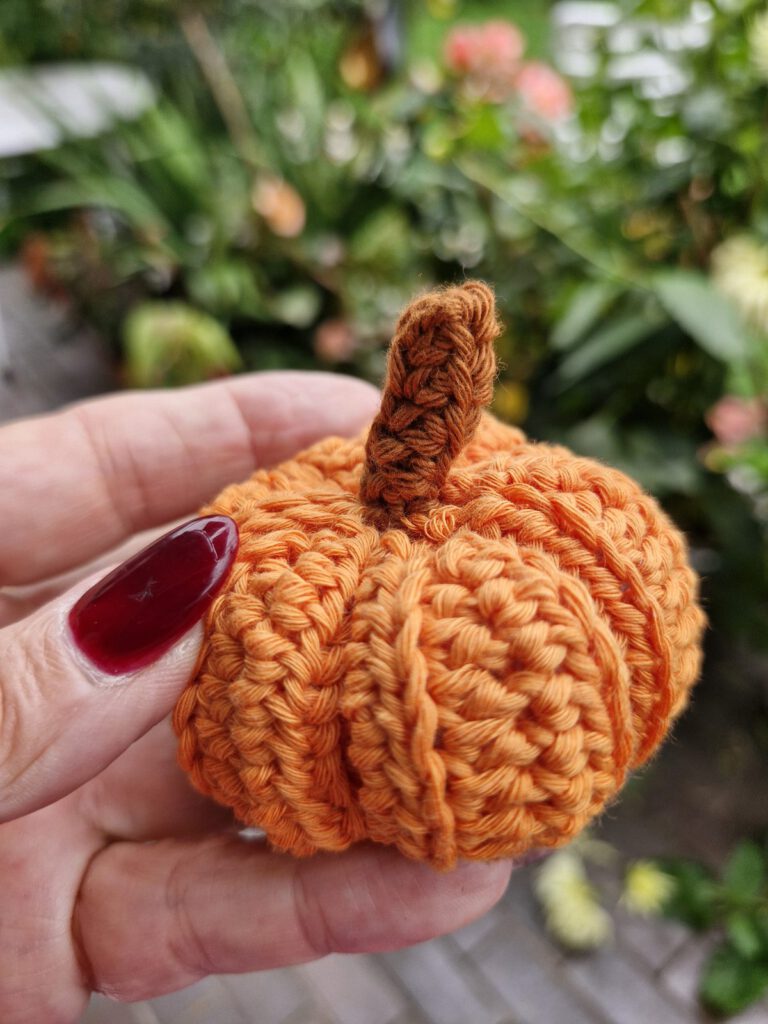 Hand holding a small orange cotton crochet pumpkin with a brown stem
