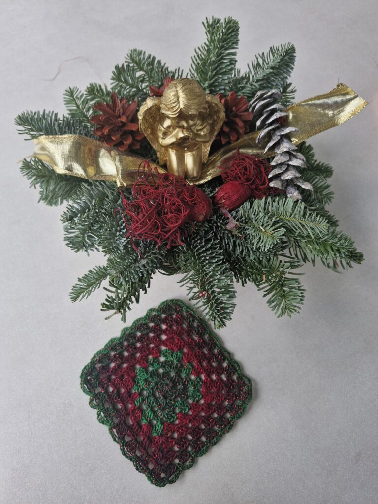 Crocheted glitter granny square in red and green next to a Christmas arrangement with a golden angel