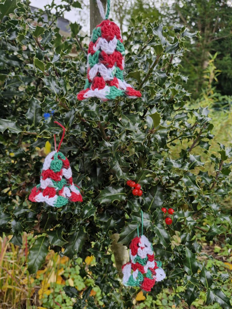 Three crocheted red, green and white Christmas bell ornaments hanging in a holly bush